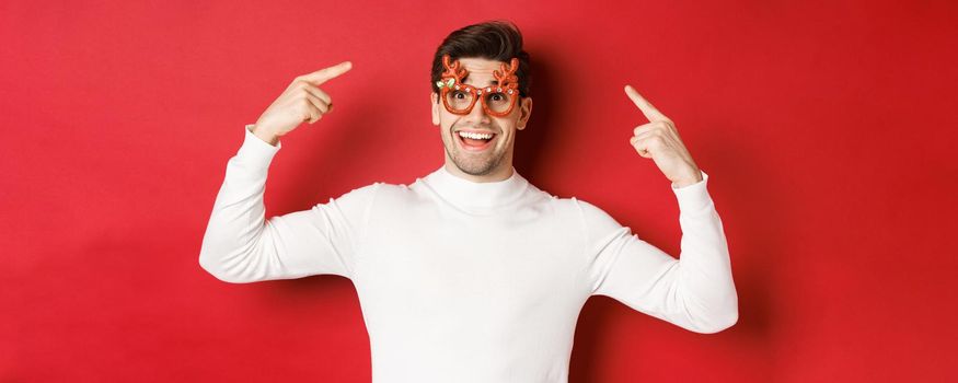 Handsome happy guy in white sweater, pointing at his christmas party glasses, celebrating new year and having fun, standing over red background.