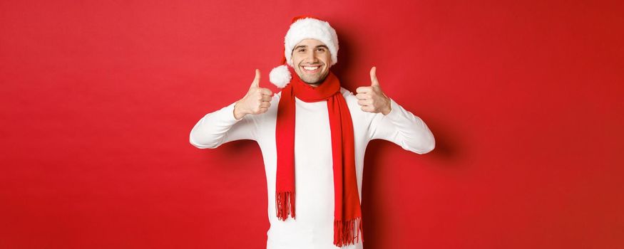 Portrait of handsome smiling man in santa scarf and hat, showing thumbs-up, celebrating christmas, standing over red background.