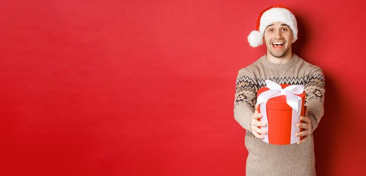 Image of happy man saying merry christmas and giving you a gift in box, smiling cheerful, celebrating winter holidays in santa hat and xmas sweater, standing over red background.