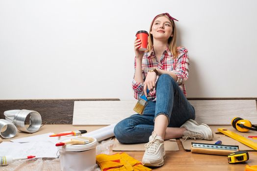 Dreamy girl painter relaxing on floor with cup of coffee. House remodeling and interior renovation concept with copy space. Young woman in red checkered shirt and jeans planning to redesign her home.