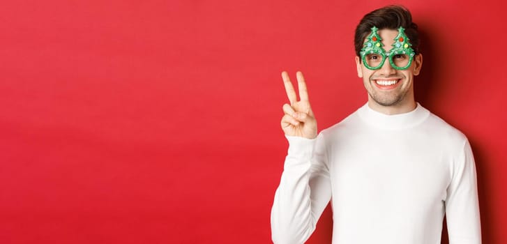 Image of handsome man in white sweater and party glasses, showing peace sign and smiling, wishing merry christmas, standing over red background.