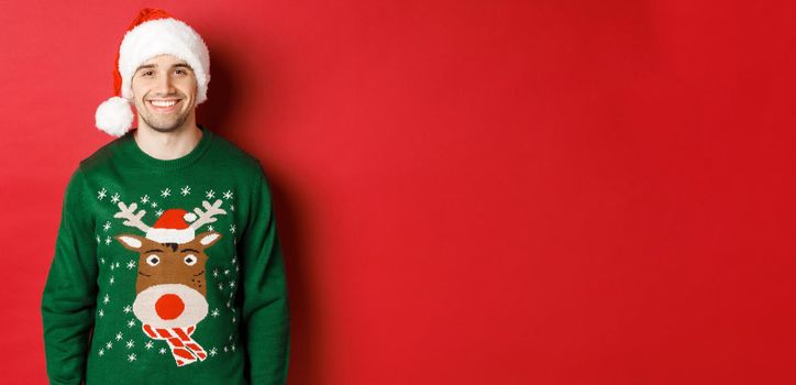 Concept of winter holidays, christmas and lifestyle. Handsome guy with bristle, wearing santa hat and green sweater, smiling joyful, celebrating new year, standing over red background.
