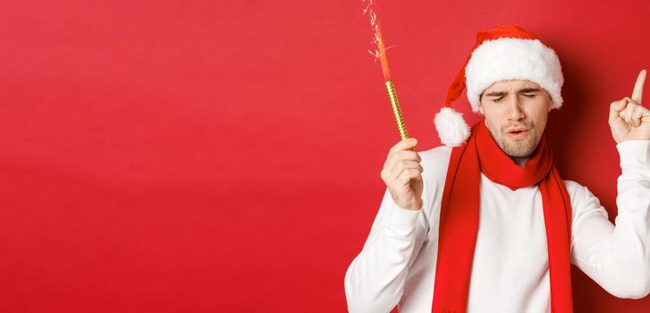 Concept of christmas, winter holidays and celebration. Attractive guy enjoying new year party, dancing with sparkler, wearing santa hat and scarf, standing over red background.