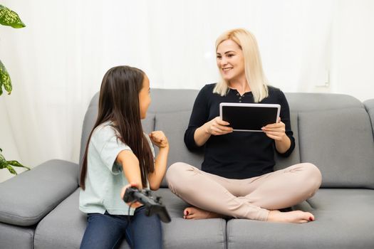 good relationship cute little girl with young mother using joystick playing video game sitting together in living room enjoying family holiday.