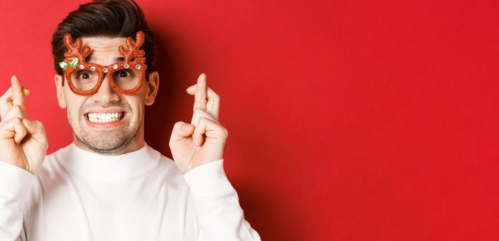 Concept of winter holidays, christmas and celebration. Close-up of hopeful man in party glasses, crossing fingers and making a wish on new year, standing over red background.