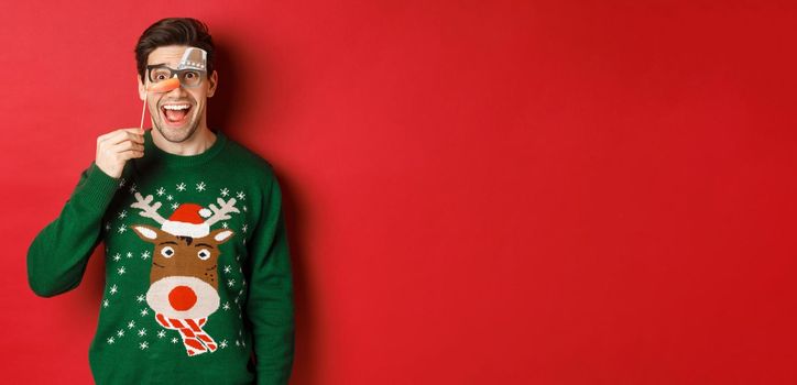 Portrait of amused handsome man in christmas sweater, holding funny party mask and smiling, celebrating winter holidays, standing over red background.