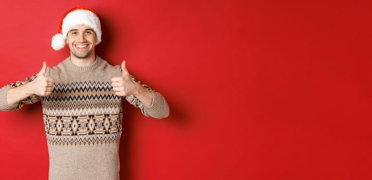 Image of cheerful attractive man in winter sweater and santa hat, showing thumbs-up, celebrating christmas and looking happy, like something, standing over red background.