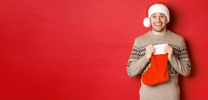 Concept of winter holidays, new year and celebration. Image of surprised and happy man receiving gift from secret santa inside christmas stocking bag, open gift and looking thankful.