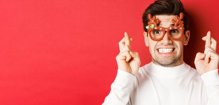 Concept of winter holidays, christmas and celebration. Close-up of hopeful man in party glasses, crossing fingers and making a wish on new year, standing over red background.