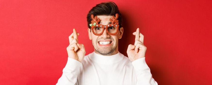 Concept of winter holidays, christmas and celebration. Close-up of hopeful man in party glasses, crossing fingers and making a wish on new year, standing over red background.