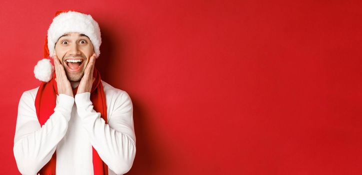 Concept of christmas, winter holidays and celebration. Close-up of surprised and happy man in santa hat and scarf, looking at something amazing, standing over red background.