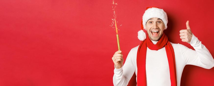 Concept of christmas, winter holidays and celebration. Handsome man celebrating new year and having fun, holding sparkler and showing thumb-up, standing over red background.