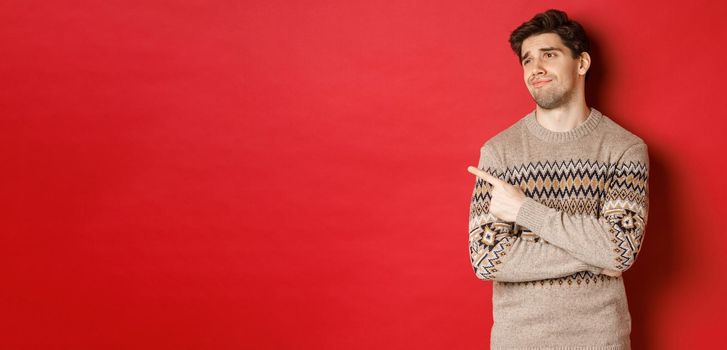 Image of disappointed handsome man in christmas sweater, looking and pointing left with upset face, express regret, showing new year advertisement, standing over red background.