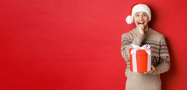 Image of handsome guy in santa hat and christmas sweater, excited to open xmas gift, looking amazed and holding present in one hand, standing over red background.