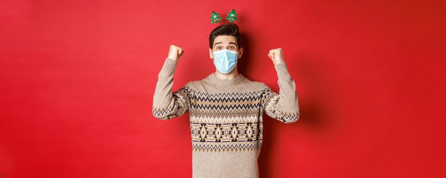 Concept of christmas, holidays and coronavirus. Happy man celebrating new year during covid-19 outbreak, wearing medical mask and sweater, rejoicing over red background.