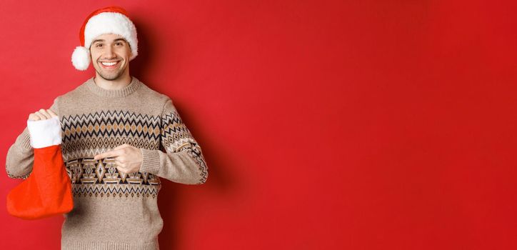 Concept of winter holidays, new year and celebration. Handsome smiling man prepared gifts for kids, pointing at christmas stocking bag, standing over red background.