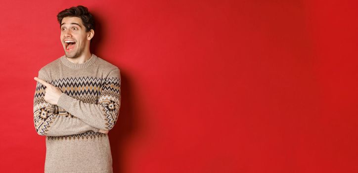 Portrait of attractive caucasian man celebrating new year, winter holidays, pointing finger and looking left with amazed expression, showing christmas advertisement, standing over red background.