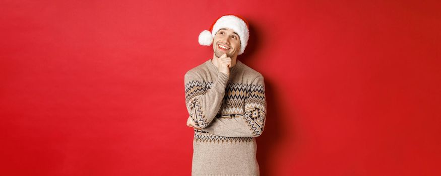 Image of happy young man in santa hat and christmas sweater, imaging something, thinking about new year gifts and smiling, looking at upper left corner, standing over red background.