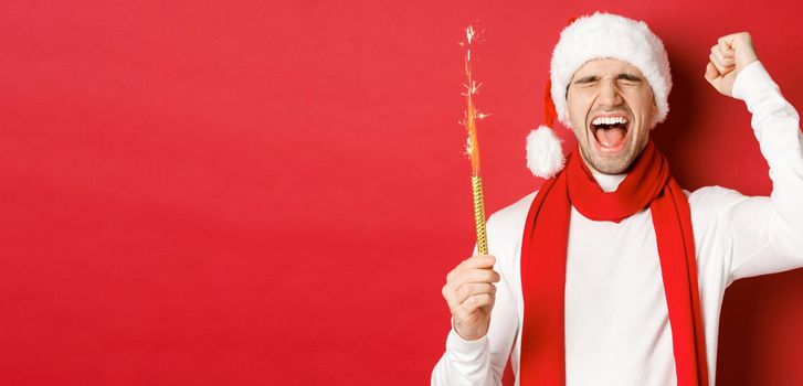 Concept of christmas, winter holidays and celebration. Handsome man celebrating new year and having fun, holding sparkler and smiling, wearing santa hat, standing over red background.