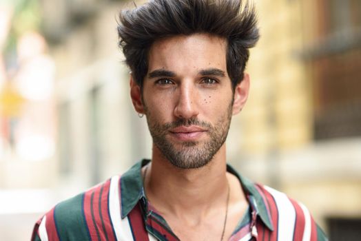 Handsome young man with dark hair and modern hairstyle wearing casual clothes in urban background.