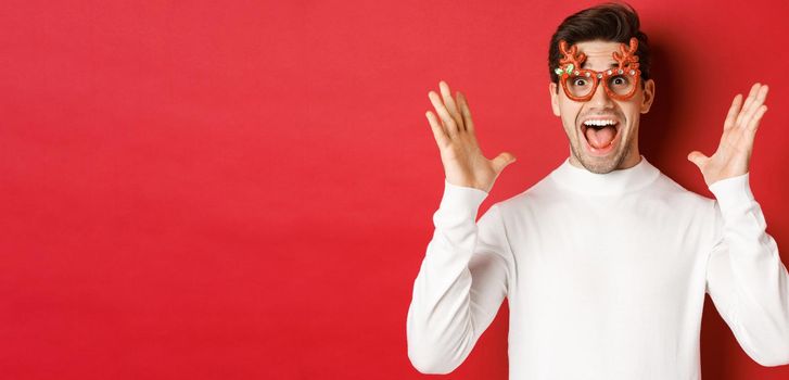 Close-up of surprised handsome guy in white sweater and party glasses, hear amazing christmas promo offer, rejoicing and standing over red background.