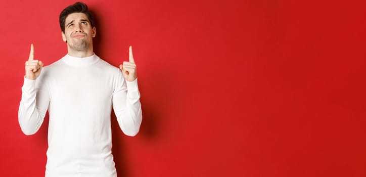 Concept of winter holidays. Doubtful handsome man in white sweater, looking skeptical and pointing fingers up at logo, standing against red background.