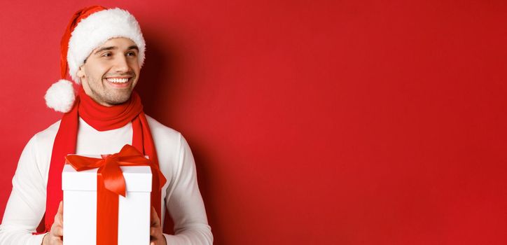 Concept of winter holidays, christmas and lifestyle. Close-up of attractive man in santa hat and scarf, holding new year gift, looking right and smiling, standing over red background.