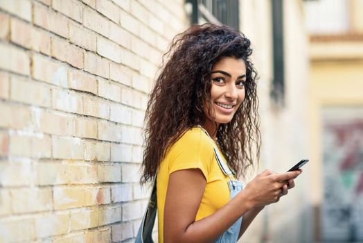 Young Arab woman texting with her smart phone on brick urban wall. Smiling North Afrincan girl in casual clothes with black curly hairstyle.