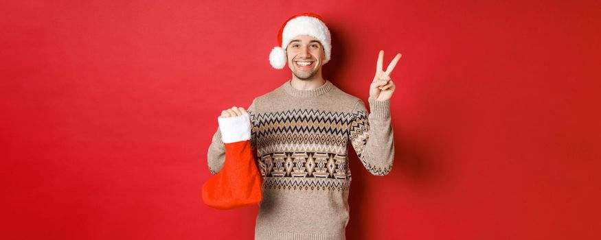 Concept of winter holidays, new year and celebration. Image of happy smiling man in santa hat and sweater, showing peace sign and a christmas stocking bag with gifts, red background.
