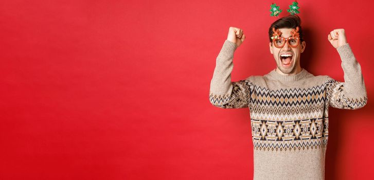 Portrait of thrilled and happy man in party glasses, winter sweater, shouting for joy and raising hands up, winning prize on christmas, achieve new year goal, standing red background.