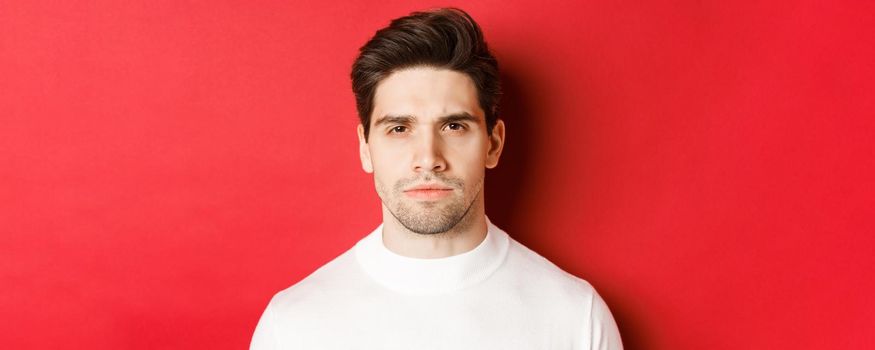 Close-up of thoughtful, serious-looking man in white sweater, standing over red background.