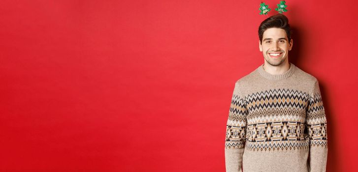 Concept of winter holidays, christmas and celebration. Handsome bearded guy in sweater wishing happy new year, smiling at camera, standing over red background.