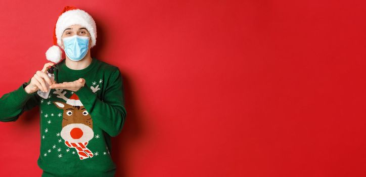 Concept of new year, coronavirus and social distancing. Attractive young man in santa hat, medical mask and christmas sweater, using hand sanitizer to clean hands, standing over red background.