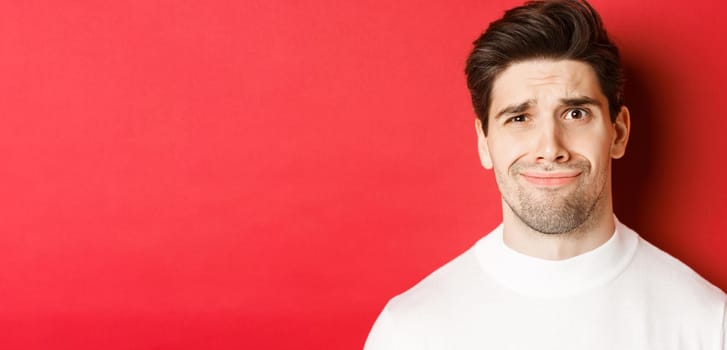 Close-up of handsome man feeling uncomfortable, grimacing and looking at something displeasing, standing over red background.