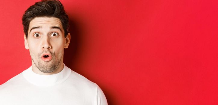 Close-up of surprised handsome man in white sweater, open mouth and looking wondered, hear amazing news, standing over red background.