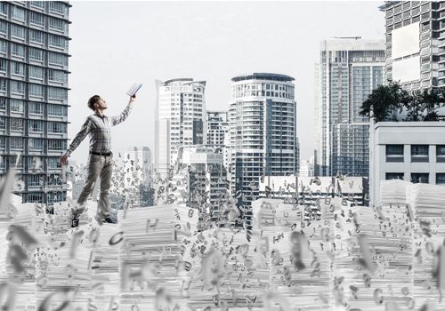 Man in casual wear keeping hand with book up while standing among flying letters with cityscape on background. Mixed media.