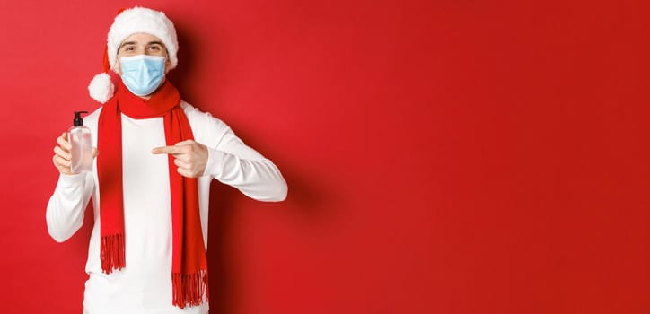 Concept of covid-19, christmas and holidays during pandemic. Handsome happy man in santa hat and medical mask, recommending use hand sanitizer, standing over red background.