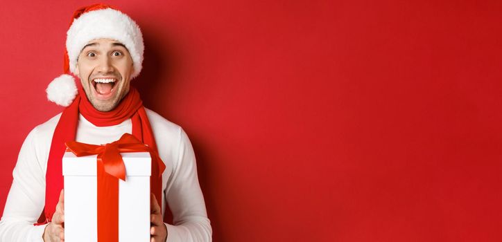 Concept of winter holidays, christmas and lifestyle. Close-up of excited handsome man in santa jat and scarf, looking amazed and receiving new year present, standing over red background.