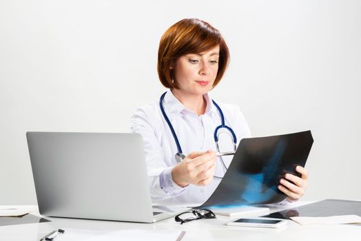 Beautiful doctor looking at x-ray scan in office. Physician in white coat with stethoscope sitting at desk with laptop. Professional medical diagnosis and treatment in clinic. Medical check up service