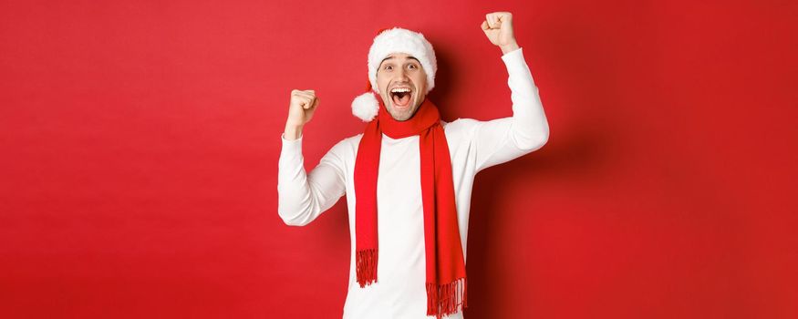 Image of joyful caucasian man in santa hat and scarf, shouting for joy and raising hands, celebrating victory or win, triumphing over red background.