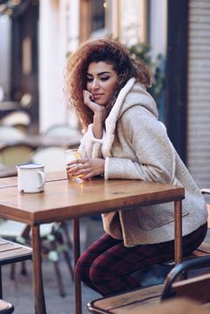 Young arabic woman sitting in an urban bar in the street. Arab girl in winter casual clothes drinking a soda outdoors.