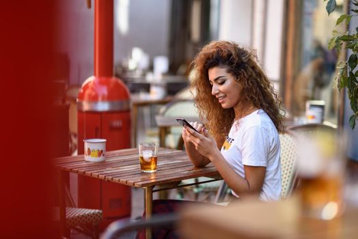 Young arabic woman smiling and sitting in an urban bar in the street looking at her smartphone. Arab girl in casual clothes drinking a soda outdoors.