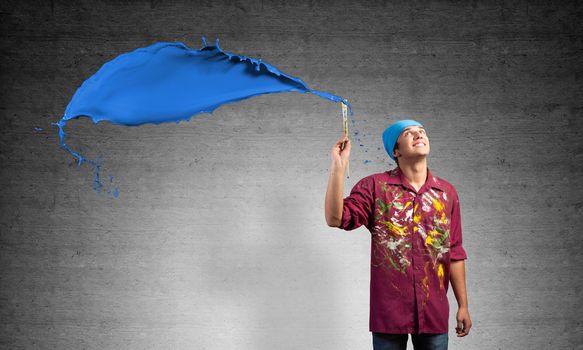 Young artist painting blue brush stroke. Male painter in dirty shirt and bandana with paintbrush on gray wall background. Creative hobby and artistic occupation. Art classes concept with copy space.