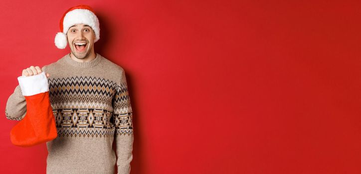 Concept of winter holidays, new year and celebration. Cheerful and surprised adult man receiving candies on Saint Nicholas day in red stocking, standing amazed over red background.