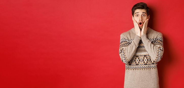 Image of surprised handsome guy reacting to cool new year promo offer, gasping amazed, wearing christmas sweater, standing over red background.