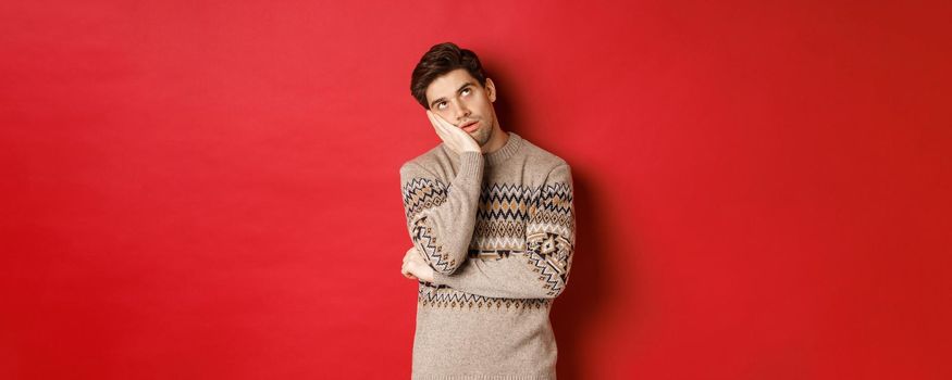 Image of annoyed young man in christmas sweater, roll eyes and leaning on hand, express displeasure and irritation, standing over red background.