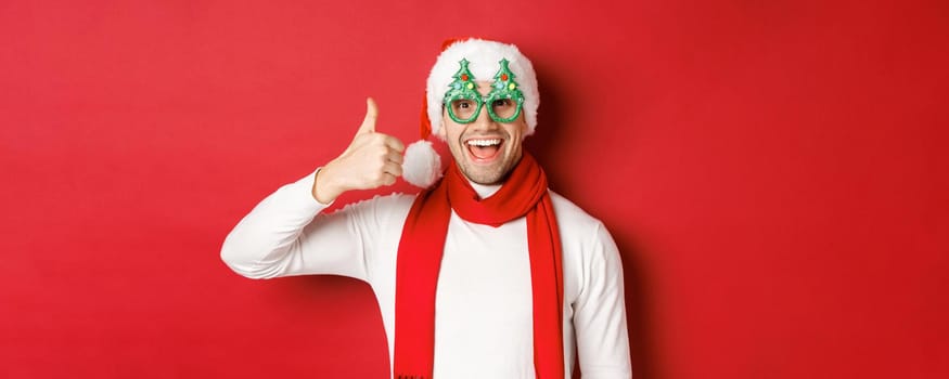 Concept of christmas, winter holidays and celebration. Cheerful guy in santa hat and party glasses, showing thumb-up and smiling pleased, standing over red background.