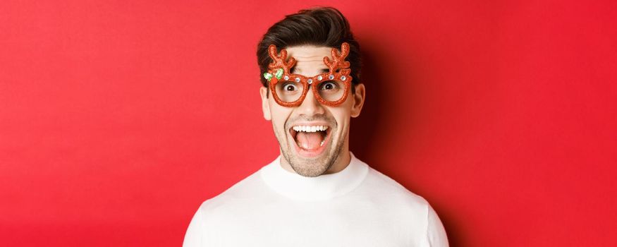 Concept of winter holidays, christmas and celebration. Close-up of attractive guy with bristle, wearing new year party glasses and looking amazed at promo offer, standing over red background.