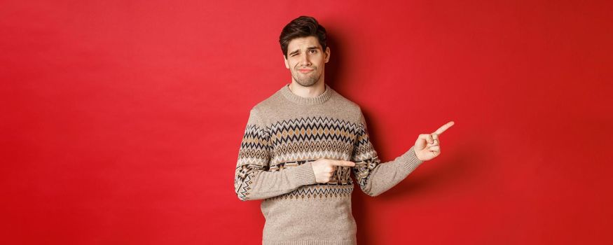 Concept of christmas celebration, winter holidays and lifestyle. Image of skeptical and displeased handsome man in xmas sweater, pointing fingers right and do not recommend product.