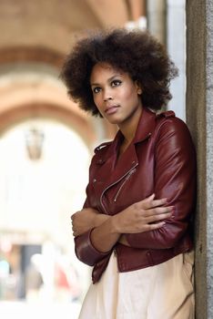 Young black female with afro hairstyle standing in an urban street. Mixed woman wearing red leather jacket and white dress with city background.